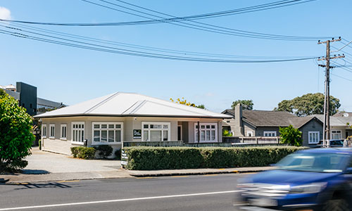 A single-storey house with a light-coloured exterior sits behind hedges on a sunny day. A blue car is driving past on the road in front. Power lines and poles are visible overhead.