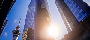 Two tall glass skyscrapers reflect sunlight with a communications tower in the background, rising high above what could be a modern PLC Header facility. The photo is taken from a low angle, looking up at the buildings and the bright sky.