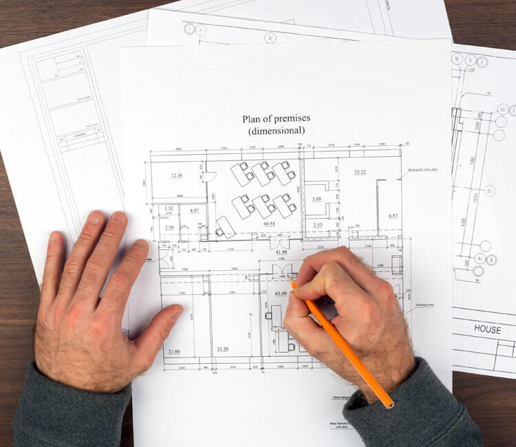 A person’s hands are working on architectural floor plans with a pencil on a wooden table. The plans, which might be required by a cross lease solicitor, show room layouts, measurements, and labelled areas such as offices and conference rooms. Other drawings are partially visible underneath.