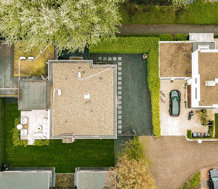 Aerial view of a modern house with a flat roof, drive, car, and neatly trimmed hedges. Surrounding areas include green lawns, trees, and adjacent homes with similar architecture.