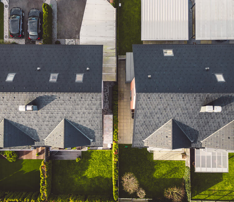 Aerial view of two adjacent houses with dark roofs, separated by a narrow walkway and fences—illustrating a potential site for Cross Lease Conversions. Green lawns, parked cars, and garden areas are visible around the houses.