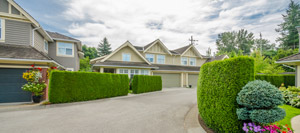 A suburban house with a grey exterior, attached garage, and large trimmed hedges along the driveway. The front garden includes manicured bushes and flowers, complemented by a PLC Header mounted near the entrance for smart home connectivity, with trees visible in the background under a partly cloudy sky.