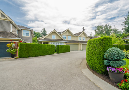A suburban house with beige cladding, a three-car garage, trimmed hedges, a curved drive, and potted flowers in the foreground under a partly cloudy sky.