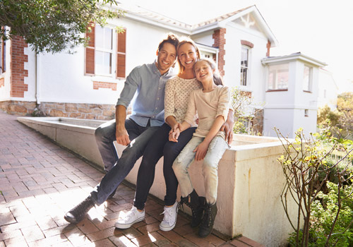 Three people, two adults and one child, sit closely together on a low wall outside a white house with brick details. They are smiling and appear to be enjoying a sunny day.