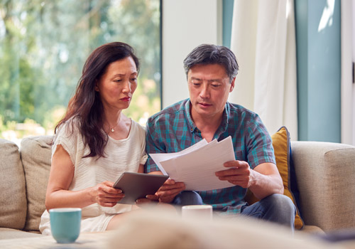 A man and woman sit on a sofa looking at papers and a tablet. They appear focused and serious. There are two mugs on the table in front of them, and a window with greenery in the background.