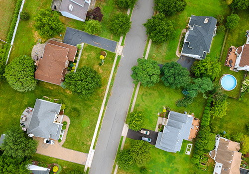 Aerial view of a suburban neighbourhood with houses, green lawns, trees, driveways, and a road running vertically through the centre. One back garden has a round swimming pool.