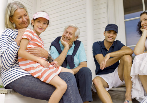 Five people sit on a veranda. An older woman holds a smiling girl on her lap. An older man sits next to them, smiling, whilst a younger man and woman sit nearby, all casually dressed and looking at each other.