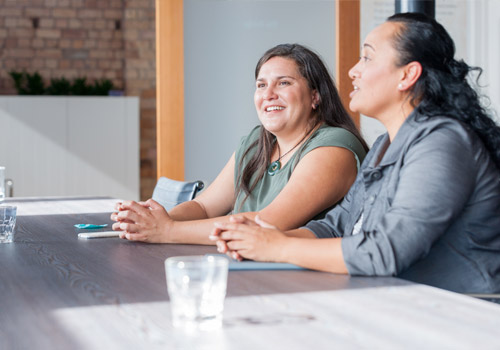 Two women are sitting at a table in a bright meeting room, engaged in conversation. Both are smiling. Water glasses and a mobile phone are on the table. The background includes a brick wall and indoor plants.