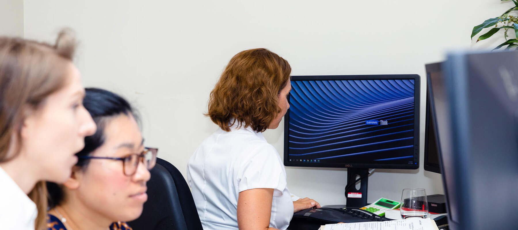 Three women sit at desks with computer monitors. One woman, seen from behind, is focused on her screen displaying a blue abstract pattern—perhaps analysing data related to legal service prices. Papers, a mouse, and a drink are on the desk. A plant is in the background.
