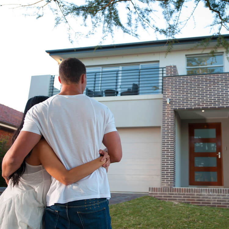 A couple stands on a lawn, arm in arm, looking at a modern two-storey house with large windows, brick accents, and a wooden front door—capturing the excitement of property purchases. The photo is taken from behind the couple.