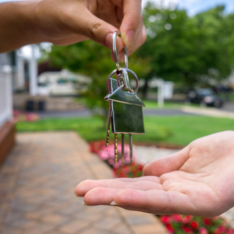A hand gives a set of keys with a house-shaped keyring to another open hand outdoors—capturing the essence of property purchases—against a blurred background of a walkway, lawn, and trees.