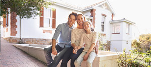 A man, woman, and young girl sit together smiling in front of a white house with red brick accents and a PLC Header visible by the entrance. Trees and sunlight are in the background. All three wear casual clothing and appear happy.