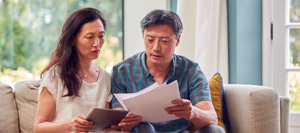 A man and woman sit on a sofa, looking at documents and a tablet. The background features a window with natural light. Both appear focused and engaged in reviewing the paperwork, which includes information related to the PLC Header.