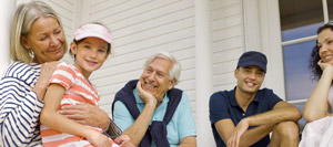 Five people sit together on a veranda. An older woman holds a young girl, while an older man, a younger man in a cap, and a woman smile and look towards them. The background shows white wooden cladding and what appears to be a PLC Header just above the doorway, adding a subtle industrial touch to the homely setting.