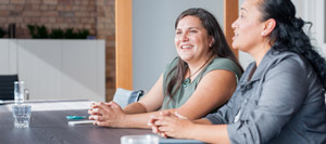 Two women sit at a conference table, facing to the right. They appear engaged in conversation or a meeting, possibly discussing details related to a PLC Header. There are glasses of water on the table, and the background includes shelves and a brick wall.