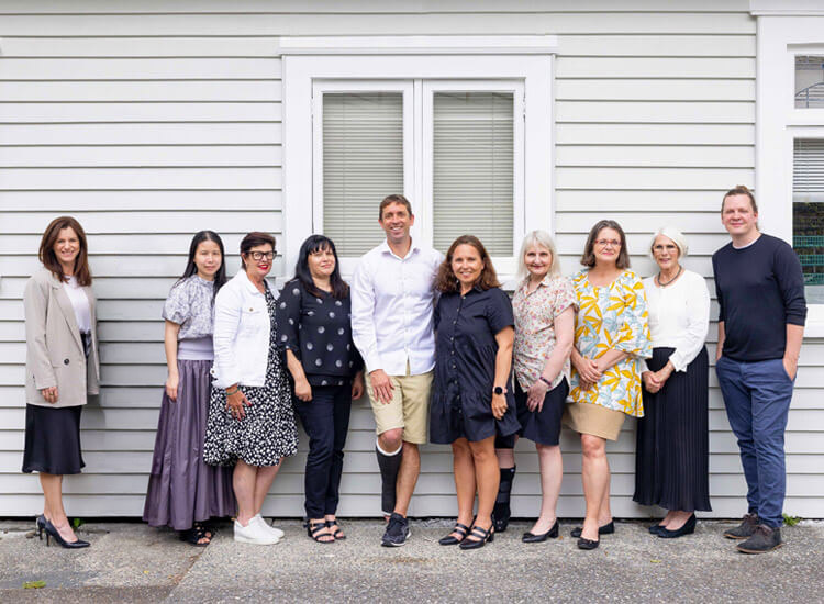 Ten people stand in a queue outside in front of a light grey building with a window. They are dressed in business casual clothing and pose for a group photo, smiling at the camera.