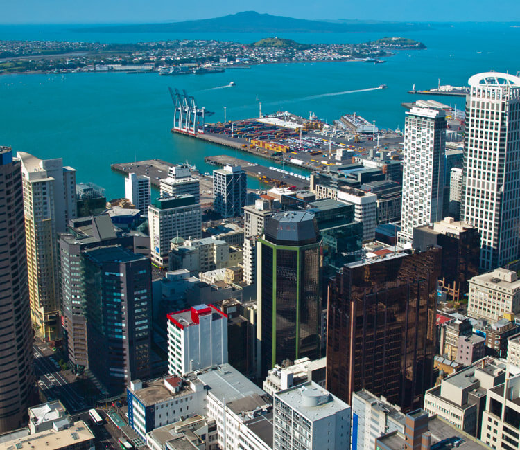 Aerial view of a city with tall buildings featuring prime commercial property, a port with cranes and containers bustling with business transactions, and blue water with an island in the background under a clear sky.