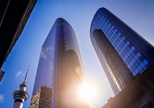 Two tall, glass-covered skyscrapers stand side by side under a blue sky. The sun is shining between them, and a communications tower with a spire is visible on the left. The buildings reflect the light and nearby surroundings.