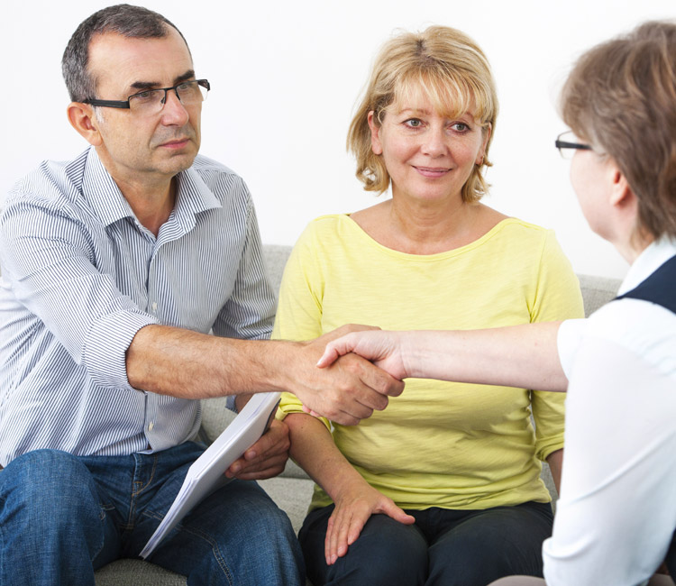 A man and a woman sit on a sofa facing another woman. The man shakes hands with her whilst holding documents related to separation agreements. The woman beside him smiles. All three appear to be having a serious conversation about relationship property.