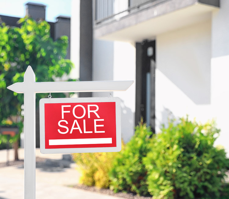 A red “For Sale” sign is hanging on a white post in front of a house with green bushes and a blurred exterior background, symbolising the changes that can follow separation agreements or the division of relationship property.