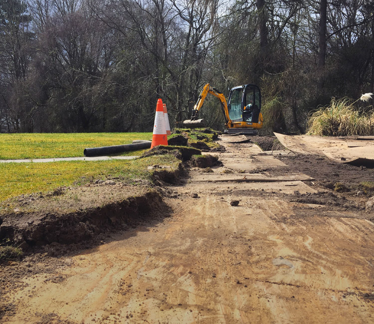 A building site with a digger, a traffic cone, earth piles, and a partially dug-up pathway—typical activity for developing new housing estates. Trees are in the background and some building materials are visible on the grass.
