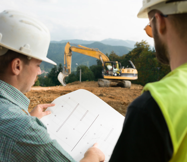 Two construction workers in hard hats examine plans at a building site. An excavator operates in the background with trees and hills visible in the distance. The focus is on the plan in the workers’ hands as they discuss details for upcoming housing estates.
