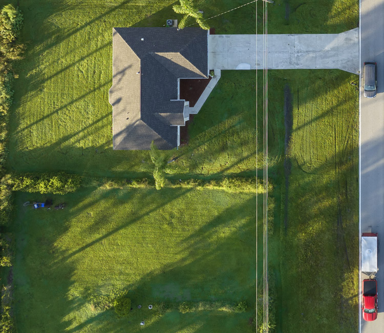 Aerial view of a house in a quiet estate, with a drive leading to the road. Two vehicles are parked on the street. The garden is green with long shadows from trees and utility wires visible across the lawn.