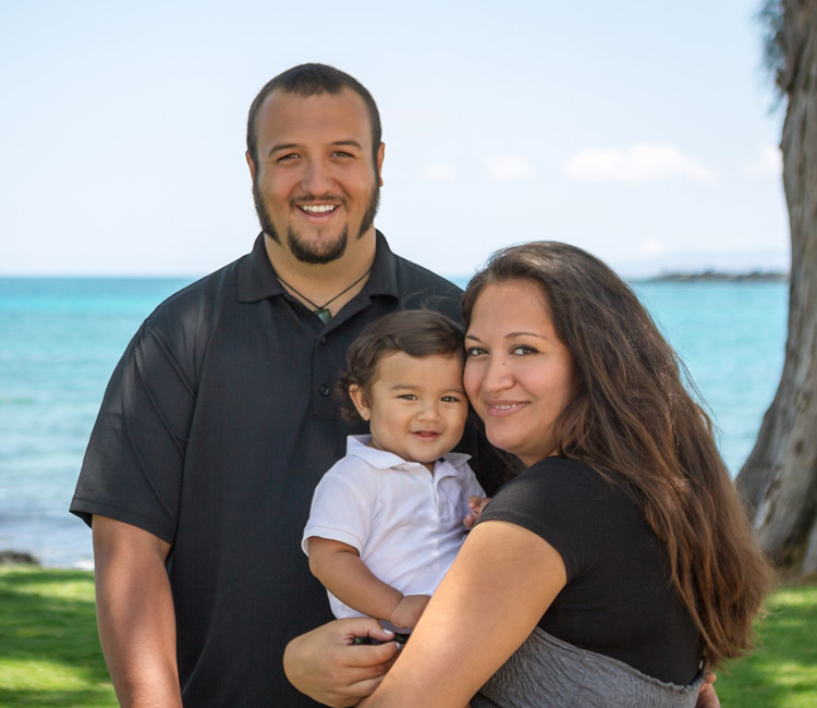 A man and woman stand outdoors near the sea, smiling at the camera while holding a young child. The sun is shining, and there is green grass and blue water in the background—a perfect moment that reminds us how important it is to plan for estate matters, ensuring your family’s future remains as bright as this day.