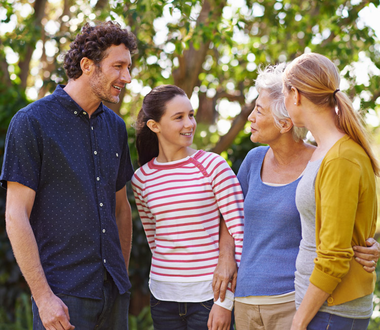 Four people stand outside in a garden or park, smiling and talking. The group includes an older woman, a younger girl, and two adults—one man and one woman—enjoying one another’s company. Surrounded by trees and greenery in the background, their warm interaction reflects strong bonds of trust between generations.
