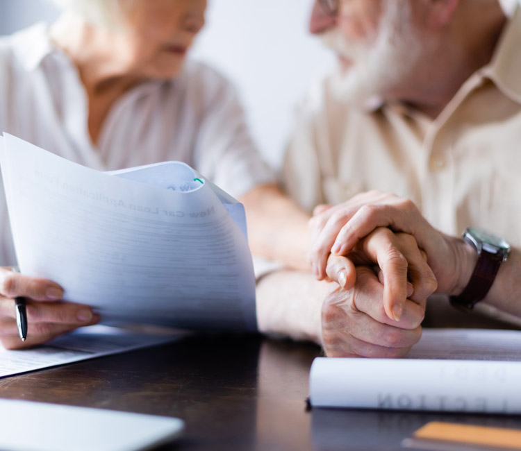 Two older adults sit together at a table, holding hands. One person holds a pen and some paperwork—possibly related to wills or Lasting Powers of Attorney—while the other rests their hand on top. Documents and a notebook are visible on the table.