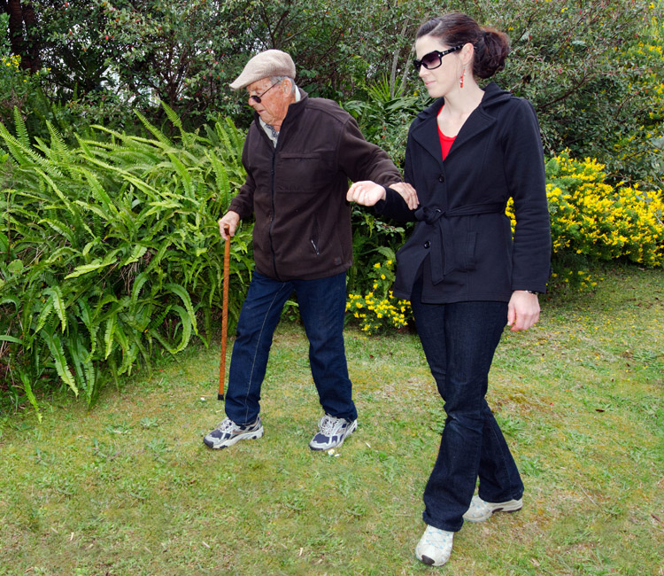 An elderly man with a walking stick walks on grass with the help of a younger woman holding his arm, highlighting the importance of support in later life—just as preparing Wills and Lasting Powers of Attorney ensures peace of mind for families. They are outdoors near green plants and yellow flowers.