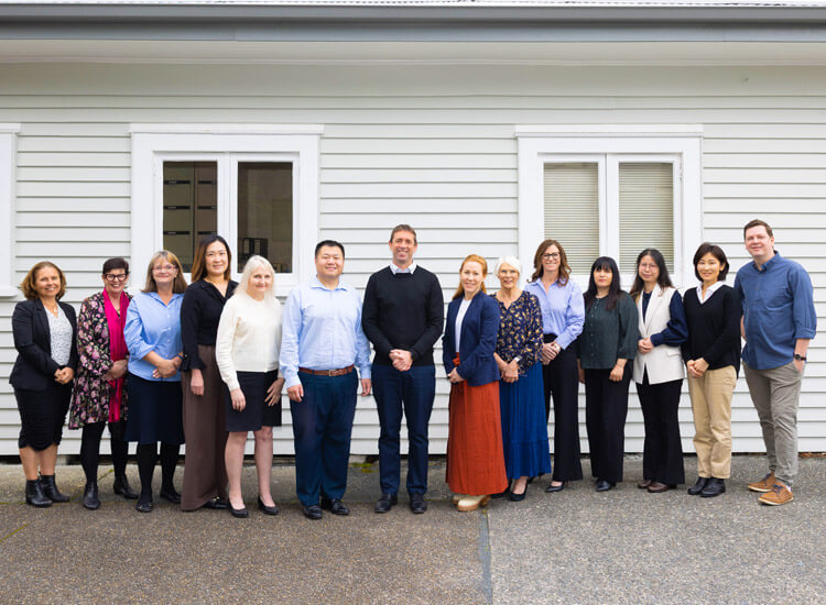 Thirteen people stand side by side outside in front of a white building with two windows, facing the camera. The group is dressed in business casual attire.