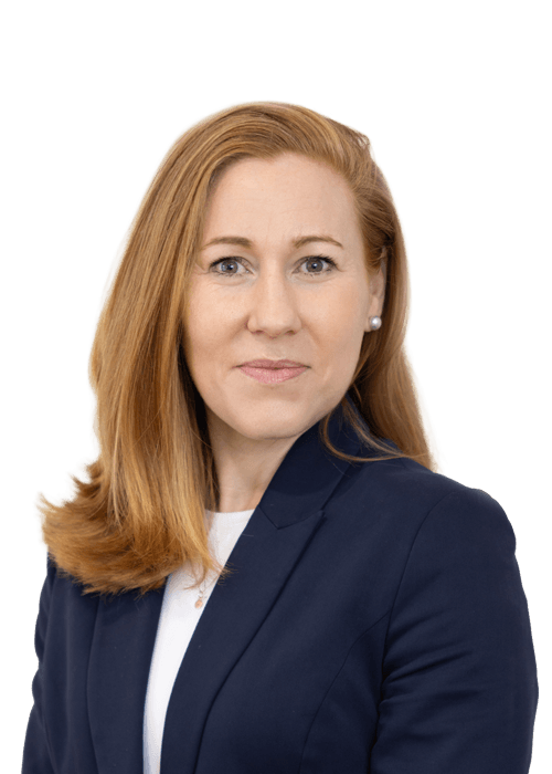A woman with straight, shoulder-length red hair wears a dark navy blazer over a white top. She is facing slightly to the side, looking directly at the camera with a neutral expression. The background is plain white.