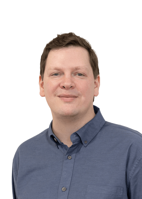 A man with short brown hair wearing a blue button-up shirt is smiling slightly while looking at the camera. The background is plain and light-coloured.
